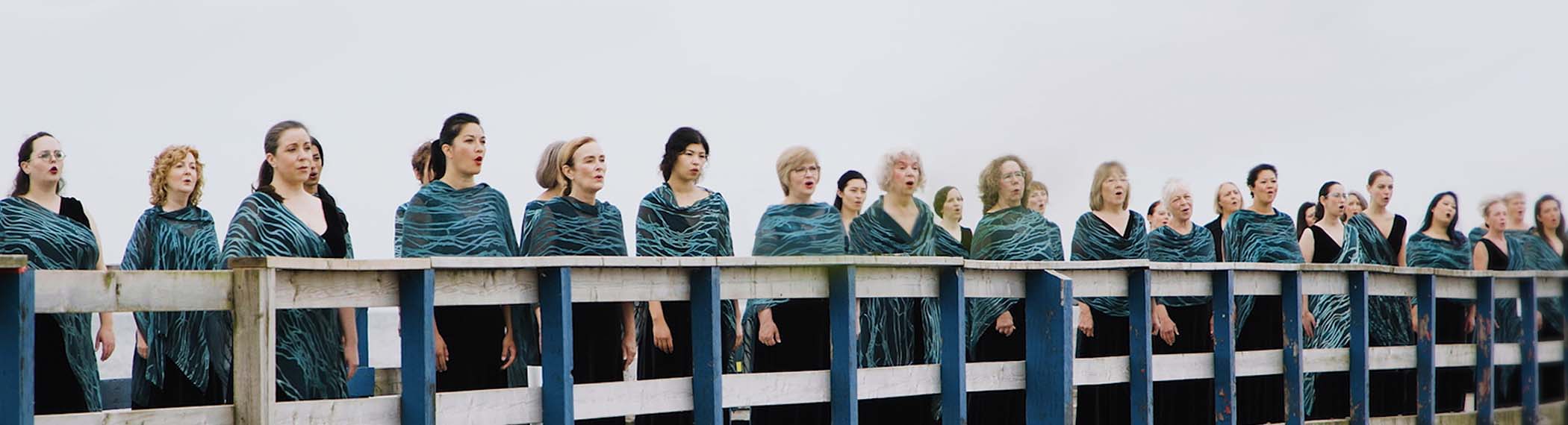 Elektra choir photograph. Choir members are standing on a bridge in black and teal uniforms, singing.